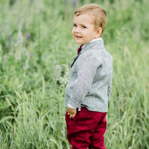 Puede incluir: Un ni&ntilde;o peque&ntilde;o con un blazer de lino gris con una corbata de lazo roja y pantalones rojos se encuentra de pie en un campo de hierba verde.