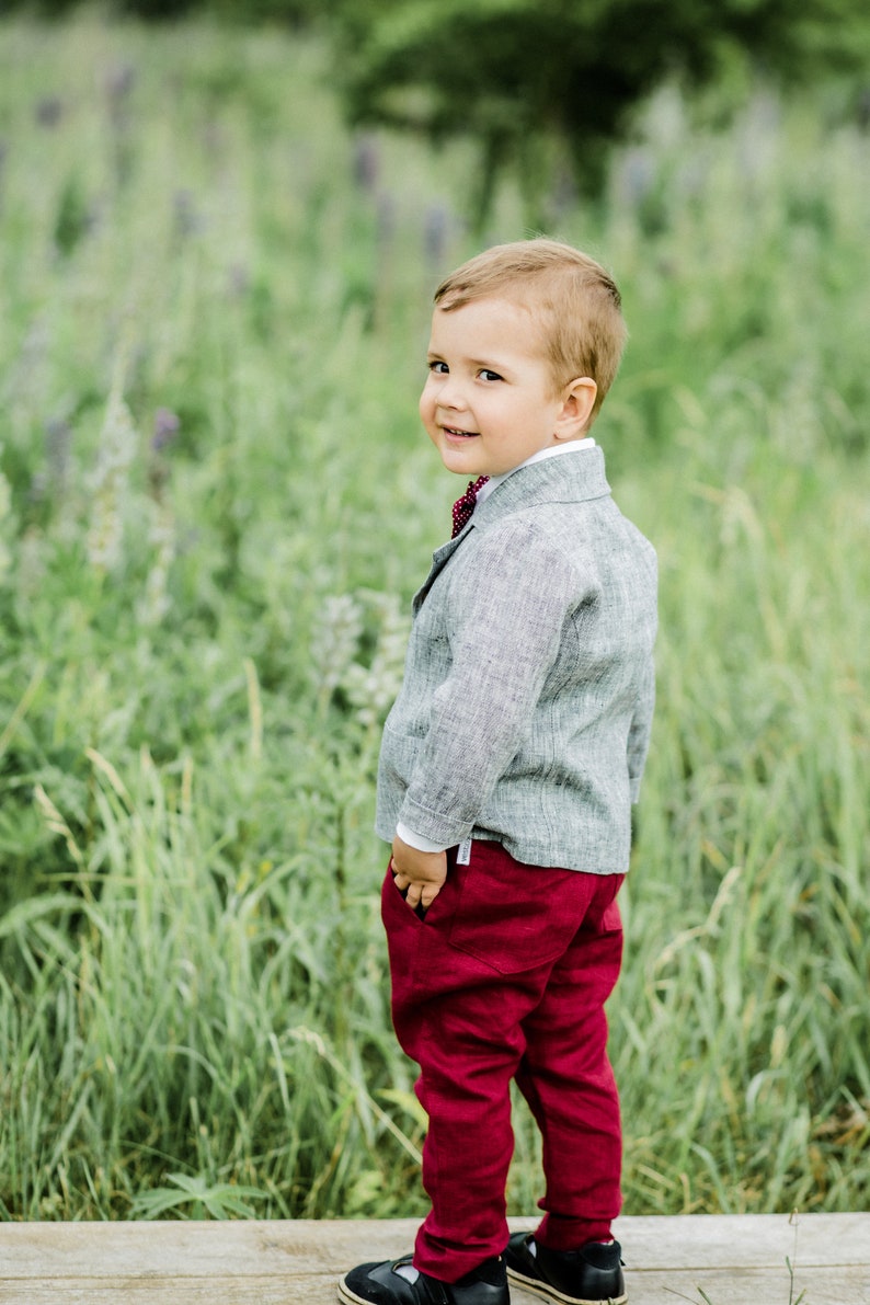 Puede incluir: Un ni&ntilde;o peque&ntilde;o con un blazer de lino gris con una corbata de lazo roja y pantalones rojos se encuentra de pie en un campo de hierba verde.