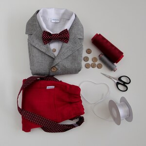 Puede incluir: Una chaqueta de lino gris con una pajarita roja, pantalones cortos rojos con lunares blancos, una bobina de hilo rojo, una bobina de hilo gris, botones de madera y una bobina de cinta blanca.
