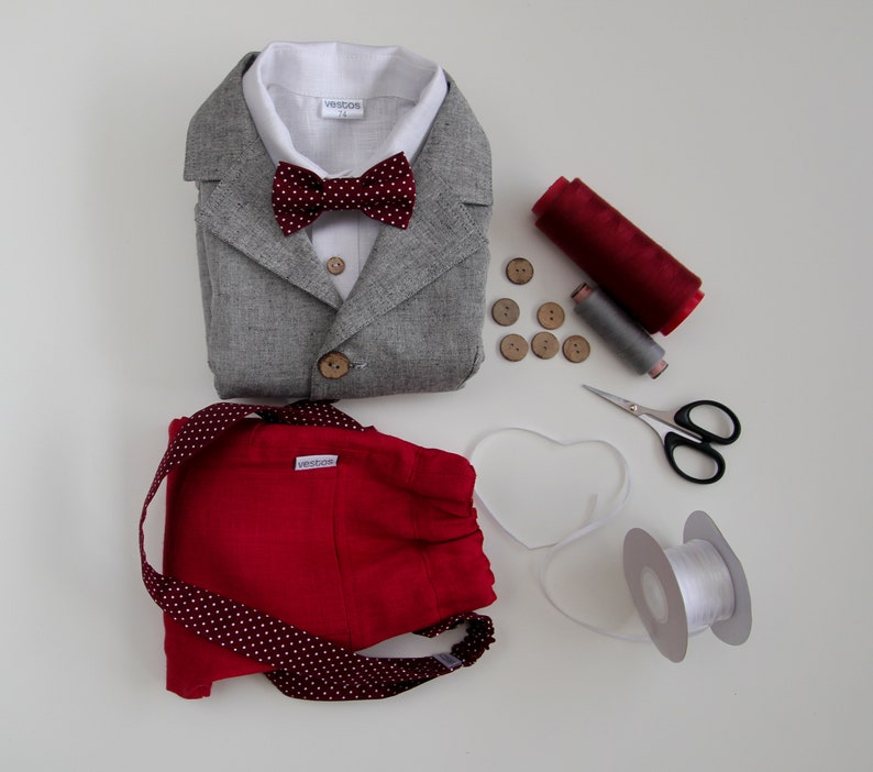 Puede incluir: Una chaqueta de lino gris con una pajarita roja, pantalones cortos rojos con lunares blancos, una bobina de hilo rojo, una bobina de hilo gris, botones de madera y una bobina de cinta blanca.