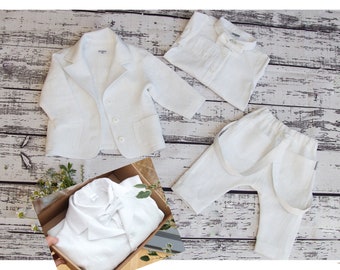 Puede incluir: Un traje de bebé de lino blanco con chaqueta, pantalón y camisa con pajarita. El traje está colocado sobre un fondo de madera blanca.