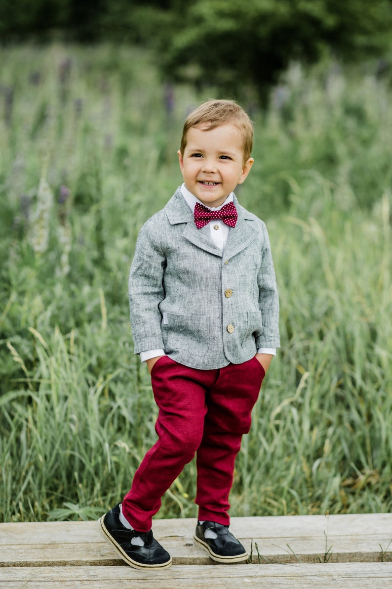 May include: A young boy wearing a gray linen blazer with a red bow tie, burgundy pants, and black shoes. He is standing on a wooden walkway in a field of green grass.