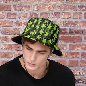 May include: A black bucket hat featuring a repeating pattern of green cannabis leaves. The hat has a black brim and is worn by a person wearing a black shirt.