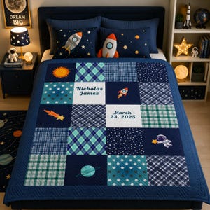 Puede incluir: Un edredón patchwork azul marino y blanco con diseños espaciales, incluyendo cohetes, estrellas y planetas. El edredón presenta el nombre "Nicholas James" y la fecha "23 de marzo de 2025". El edredón está sobre una cama con almohadas a juego.