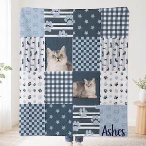Könnte beinhalten: Eine blau-weiße Patchwork-Decke mit wiederkehrenden Mustern aus Katzengesichtern, Pfotenabdrücken, Sternen, Streifen und Karomustern. Zwei Felder zeigen ein realistisches Foto einer grauen getigerten Katze. Der Name Ashes ist aufgedruckt.