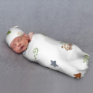 May include: A newborn baby swaddled in a white blanket and matching hat, both adorned with watercolor illustrations of a train, stars, and foliage. The baby is lying on a gray surface.
