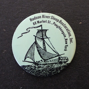 May include: A white and blue round button with a black and white illustration of a sailboat on the water. The text on the button reads "Hudson River Sloop Restoration, Inc. 88 Market St., Poughkeepsie, New York."
