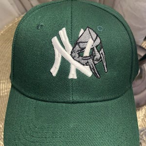 Könnte beinhalten: Eine waldgrüne Baseballkappe mit einem gestickten Logo auf der Vorderseite. Das Logo besteht aus einem großen weißen Buchstaben N, der mit einer grauen und schwarzen Maskengrafik im Metallic-Stil verschlungen ist und ein Design in der Mitte bildet.