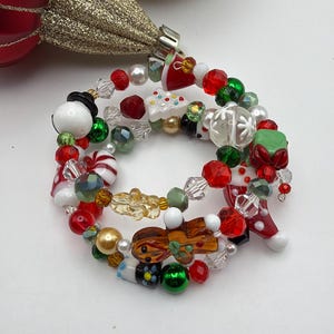 Peut inclure: Un bracelet en fil à mémoire de forme multi-rangs composé de perles sur le thème de Noël, incluant un bonhomme en pain d'épices, un bonhomme de neige, un bonbon menthe, un cadeau vert et un sapin blanc, avec des perles rouges, vertes et dorées.