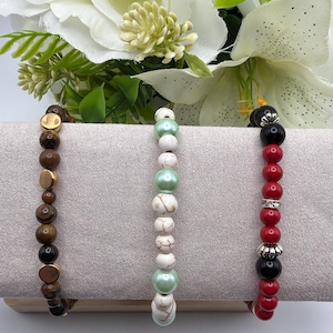 May include: Three beaded bracelets in different styles. One has brown and gold beads, another has white and mint green beads, and the third has red and black beads with a silver accent. The bracelets are displayed on a neutral surface.