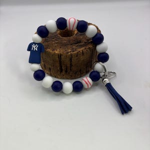 May include: A beaded keychain bracelet featuring navy blue, white, and baseball-themed beads. Includes a navy blue tassel and a small charm of a baseball jersey with the NY logo. The bracelet is designed to be worn on the wrist.