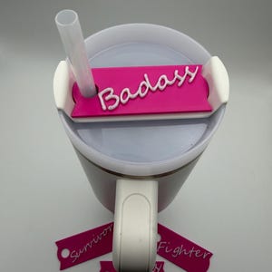 May include: A white and pink tumbler with a straw and a pink tag that says "Badass" in white script. Additional pink tags with the words "Survivor" and "Fighter" are also present.