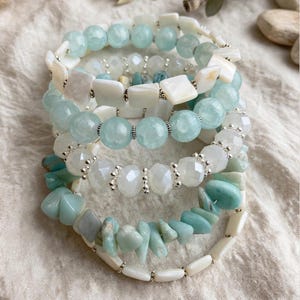 May include: A stack of five bracelets featuring a variety of light blue and white beads. The collection includes round faceted light blue stones, smooth white rectangular shell-like pieces, clear faceted crystal beads, and irregular light blue stone chips.