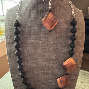 Könnte beinhalten: Ein Halsketten- und Ohrring-Set, das auf einer Schaufensterpuppe präsentiert wird. Die Halskette hat dunkelgraue Lavasteinperlen und quadratische, orange-braune Steinakzente. Die Ohrringe haben passende orange-braune quadratische Steine. Die Halskette ist silberfarben.