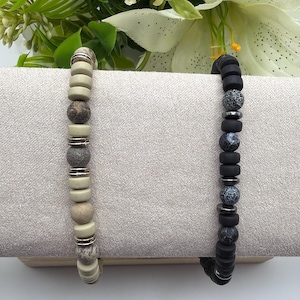 Op de afbeelding: Twee armbanden met kralen. Eén armband heeft crèmekleurige en gespikkelde kralen met zilverkleurige afstandhouders. De andere armband heeft zwarte kralen met grijs en zwart gekleurde kralen en zilverkleurige afstandhouders. De armbanden worden op een neutrale ondergrond gepresenteerd.