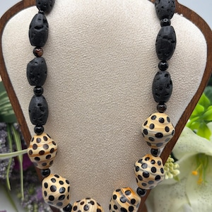 May include: A statement necklace with a striking contrast of textures. It features black lava rock beads and patterned beads with a light brown base and black polka dots. The necklace is displayed on a wooden stand, highlighting its unique design and natural materials.