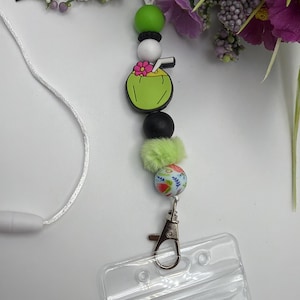 Peut inclure: Un cordon de cou à perles avec un porte-badge en plastique transparent. Le cordon comprend un charm en forme de noix de coco verte avec une fleur rose, des perles noires et blanches et une perle verte duveteuse. Un cordon blanc et un fermoir en métal complètent le design.