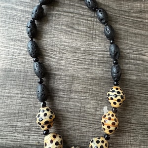 Puede incluir: Un collar con cuentas de roca volcánica negra y cuentas color crema con lunares negros. El collar tiene un cierre plateado. Las cuentas están ensartadas con un hilo negro. El collar se exhibe sobre una superficie de madera.