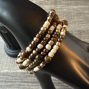 Puede incluir: Una pulsera de cuentas de varios hilos en tonos marrón, dorado y crema. La pulsera presenta pequeñas cuentas redondas y cuentas más grandes y texturizadas. La pulsera se muestra en una mano de maniquí negra.