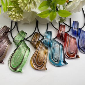 Peut inclure: Une collection de six pendentifs en verre aux tons violet, vert, ambre, turquoise, rouge et bleu, ornés de tourbillons de paillettes métalliques en forme de goutte. Chaque pendentif est suspendu à un cordon noir, offrant une finition lisse et brillante.