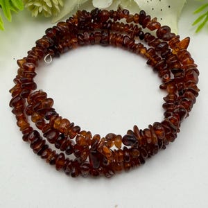 May include: A multi-strand wrap bracelet composed of numerous small, polished, irregular amber-colored resin chips. The deep brown and translucent orange beads are strung together in a continuous circular pattern, creating a layered appearance.