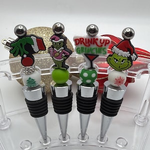May include: Four silver-toned metal bottle stoppers with black rubber rings, each topped with a decorative bead and a character charm. The charms include a Grinch hand, a Grinch in a pink outfit, a sign reading DRINK UP GRINCHES, and a Grinch face.