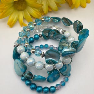 May include: A collection of turquoise and white beaded bracelets with silver details. The bracelets incorporate diverse bead shapes and sizes, including round, oval, and faceted beads. Yellow flowers are visible in the background.