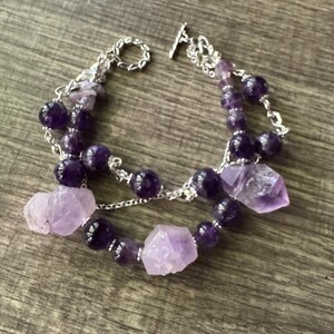 Peut inclure: Un bracelet à deux rangs avec des perles d'améthyste et des cristaux d'améthyste bruts. Le bracelet est relié par une chaîne argentée et possède un fermoir à bascule. Les pierres d'améthyste varient du lavande clair au violet foncé.