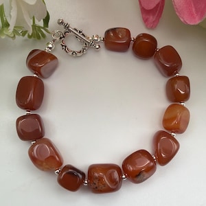 Peut inclure: Un bracelet composé de pierres d'agate polies, brun rougeâtre. Les pierres sont rectangulaires et enfilées avec des perles argentées et un fermoir à bascule. Le bracelet est présenté sur une surface blanche avec des fleurs en arrière-plan.