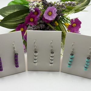 Puede incluir: Tres pares de pendientes colgantes en tarjetas de exhibición blancas. Los pendientes presentan cuentas apiladas y coloridas en púrpura, blanco y turquesa, con ganchos plateados. Un ramo de flores moradas y vegetación está en el fondo.