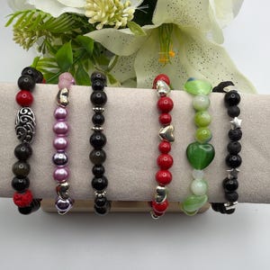 May include: Six beaded bracelets in various styles and colors. The bracelets feature beads in red, black, pink, green, and white, with silver-toned accents. Some bracelets have heart, star, and decorative bead designs. The bracelets are displayed on a neutral-colored surface.