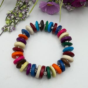 Peut inclure: Un bracelet coloré composé de perles rondes et plates en bois de différentes couleurs, dont le rouge, l'orange, le bleu et le blanc. Les perles sont enfilées avec un matériau argenté, créant un accessoire vibrant et accrocheur. Le bracelet est un bijou.