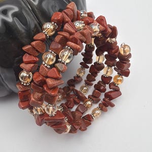 May include: A spiral bracelet featuring reddish-brown stone chips and clear, gold-toned beads. The bracelet is made of multiple strands of the stone chips and beads, creating a layered effect. The beads are interspersed throughout the design, adding a touch of sparkle.