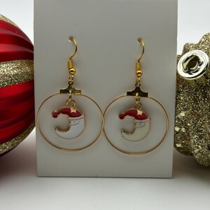Puede incluir: Pendientes de aro dorados con una luna creciente blanca que lleva un gorro de Papá Noel rojo. Los pendientes cuelgan de ganchos dorados. Un accesorio festivo para las fiestas.