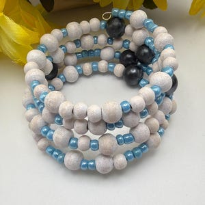 Peut inclure: Un bracelet enroulé composé de perles en bois beige clair, entrecoupées de petites perles bleu clair. Orné de perles noires. Le bracelet est enroulé en plusieurs couches, créant un effet superposé. Le design est posé sur un fond flou.