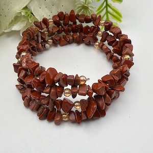 Peut inclure: Un bracelet multi-tours composé de fragments de pierre de jaspe rouge irréguliers et polis, alternés avec de petites perles rondes métalliques dorées. Le bracelet est conçu avec un fil métallique flexible en forme de spirale.