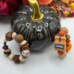 Puede incluir: Una calabaza de cerámica negra con un tallo dorado, rodeada de follaje otoñal y dos pulseras de cuentas. Una pulsera presenta abalorios con temática de café, incluyendo una cafetera y una bolsa con la etiqueta "Coffee". La otra pulsera tiene cuentas geométricas y estampadas.