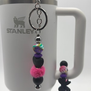 May include: A silver keyring with a beaded charm featuring a mix of colourful beads, including a pink fluffy ball, black beads, and purple accents. The keyring is next to a white Stanley mug.