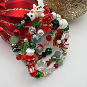 May include: A multi-strand memory wire bracelet featuring a variety of holiday-themed beads, including white snowman figures, red and white polka-dot mittens, peppermint candies, green and red faceted crystals, and clear snowflake-patterned beads.