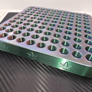 May include: A square, iridescent, multi-colored storage tray featuring a grid of circular holes. The side edges of the tray are embossed with a repeating leaf pattern. The surface displays a shifting gradient of green, blue, and purple hues.