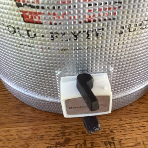 May include: A silver metal cooler with a black and white spigot. The cooler has a textured surface and the spigot has the word "Hamilton" printed on it.