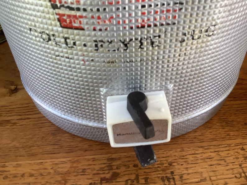 May include: A silver metal cooler with a black and white spigot. The cooler has a textured surface and the spigot has the word "Hamilton" printed on it.