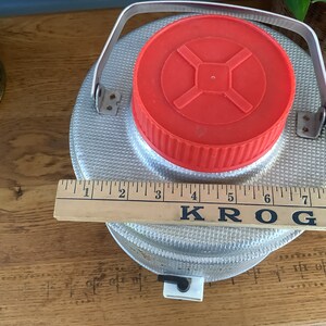 May include: A silver, metal water jug with a red lid and a handle. The jug is sitting on a wooden surface with a ruler that says "KROGER" on it.