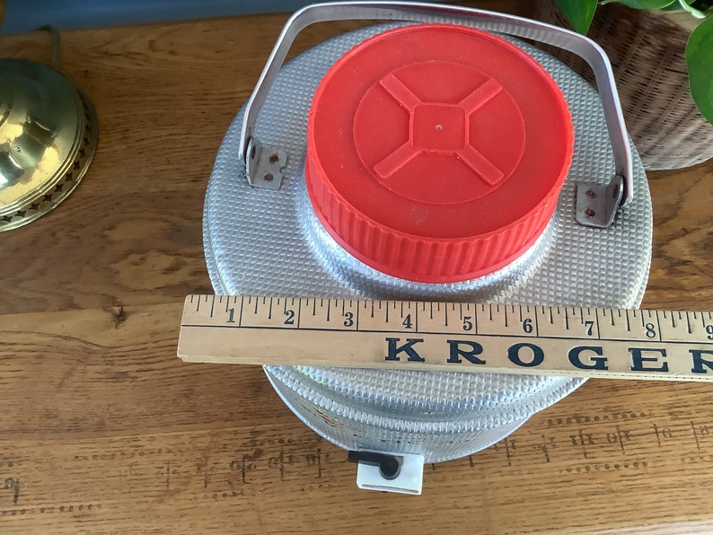 May include: A silver, metal water jug with a red lid and a handle. The jug is sitting on a wooden surface with a ruler that says "KROGER" on it.