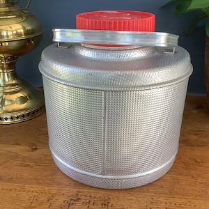 May include: A silver aluminum water jug with a red lid and a handle. The jug has a textured surface.