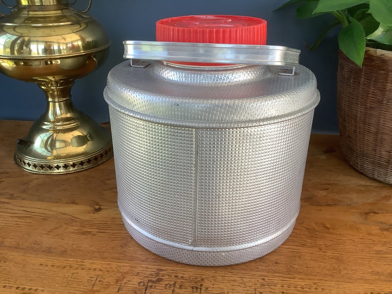 May include: A silver aluminum water jug with a red lid and a handle. The jug has a textured surface.