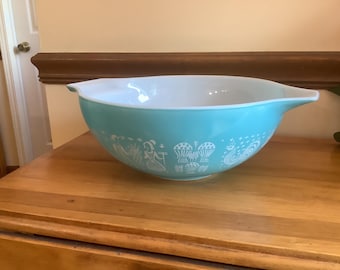 May include: A turquoise mixing bowl with a white interior and a white printed pattern featuring stylized figures, mushrooms, and a rooster. The bowl has a wide, rounded shape with a pouring spout on one side and a handle-like tab on the other side.