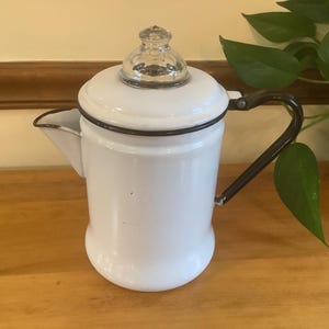 May include: White enamel coffee pot with a black handle and trim. The pot has a clear glass knob on the lid and a spout for pouring. The pot is sitting on a wooden surface.