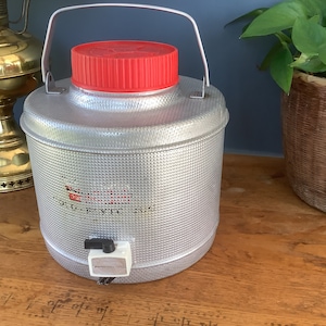May include: A silver, metal beverage dispenser with a red lid and a spigot. The dispenser has a textured surface and the words "OLD STYLE" are printed on the front.