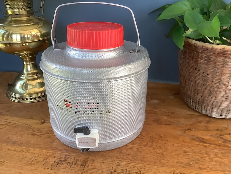 May include: A silver, metal beverage dispenser with a red lid and a spigot. The dispenser has a textured surface and the words "OLD STYLE" are printed on the front.